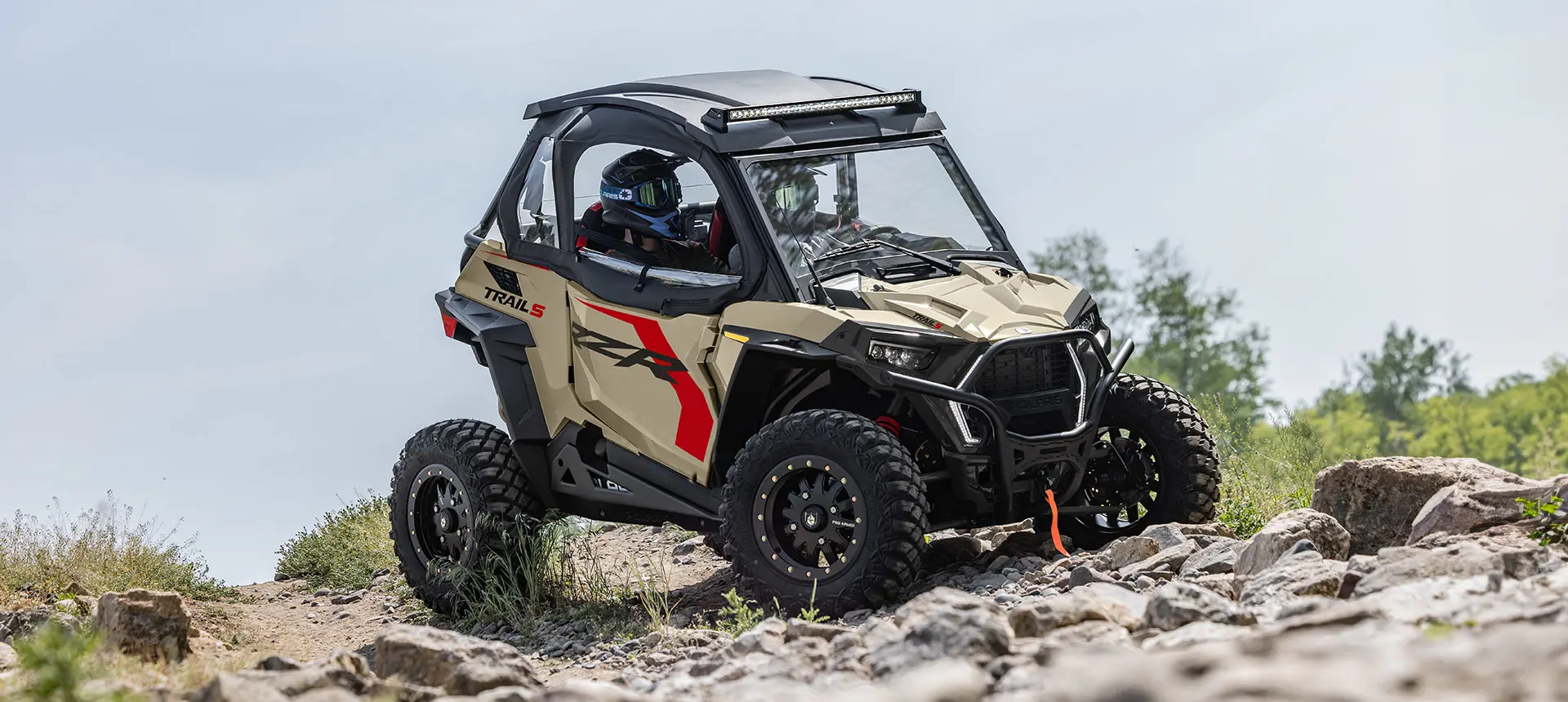 Side profile of a Polaris RZR Trail side-by-side crawling over rocky terrain.