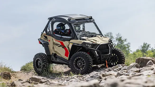 Side profile of a Polaris RZR Trail side-by-side crawling over rocky terrain.