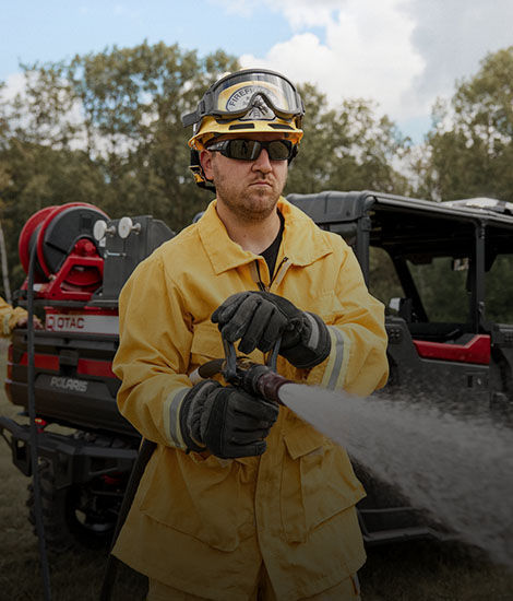 Firefighter in helmet and yellow uniform operating a Polaris Fire & Rescue UTV vehicle with a hose spraying water, outdoors with trees in the background.