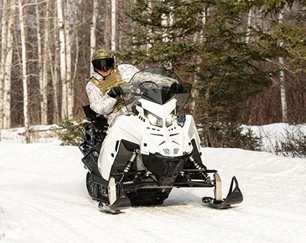 A person wearing winter camouflage gear rides a white and black snowmobile 2026 Military 850 TITAN 155 on a snowy path lined with trees.