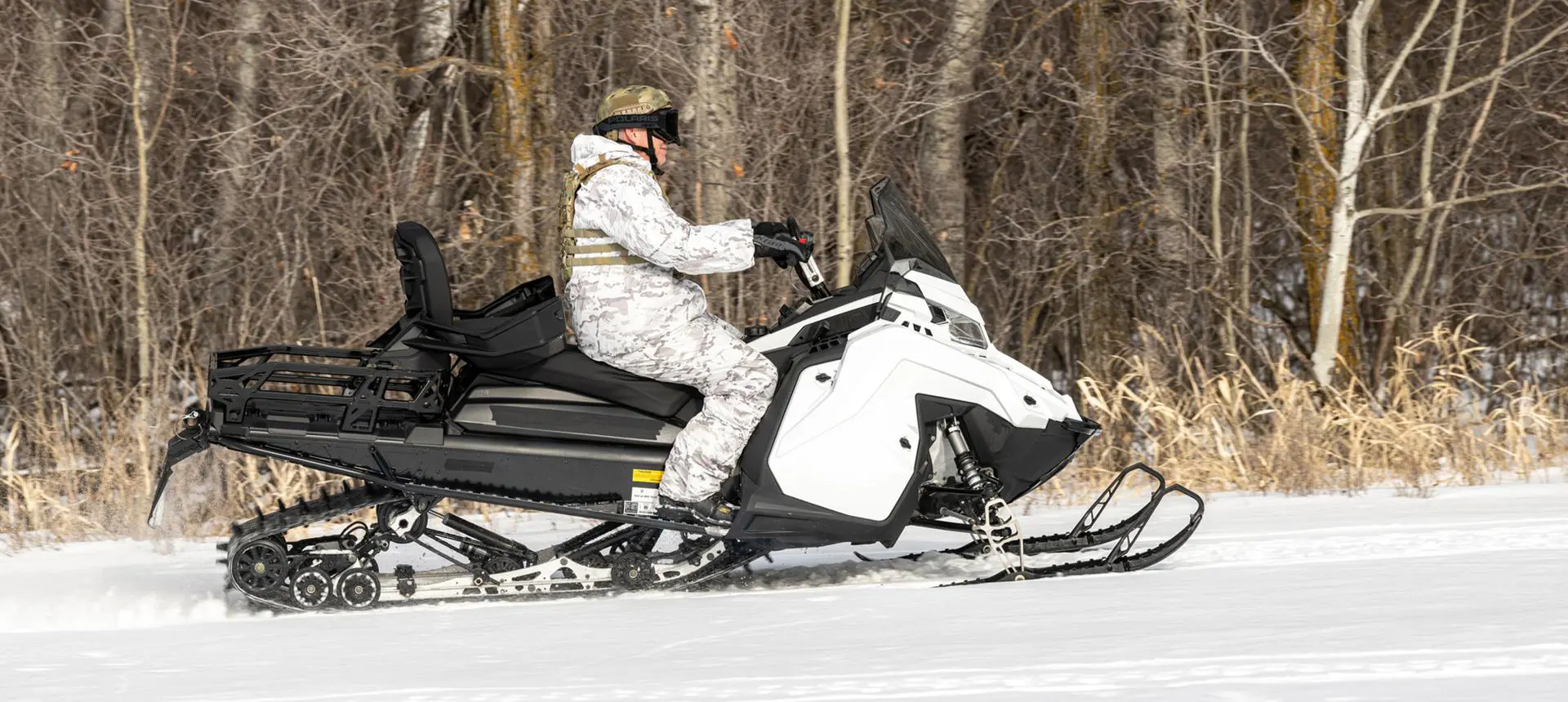 A person dressed in white camouflage drives a white and black snowmobile 2026 Military 850 TITAN 155 across a snow-covered field with trees in the background.