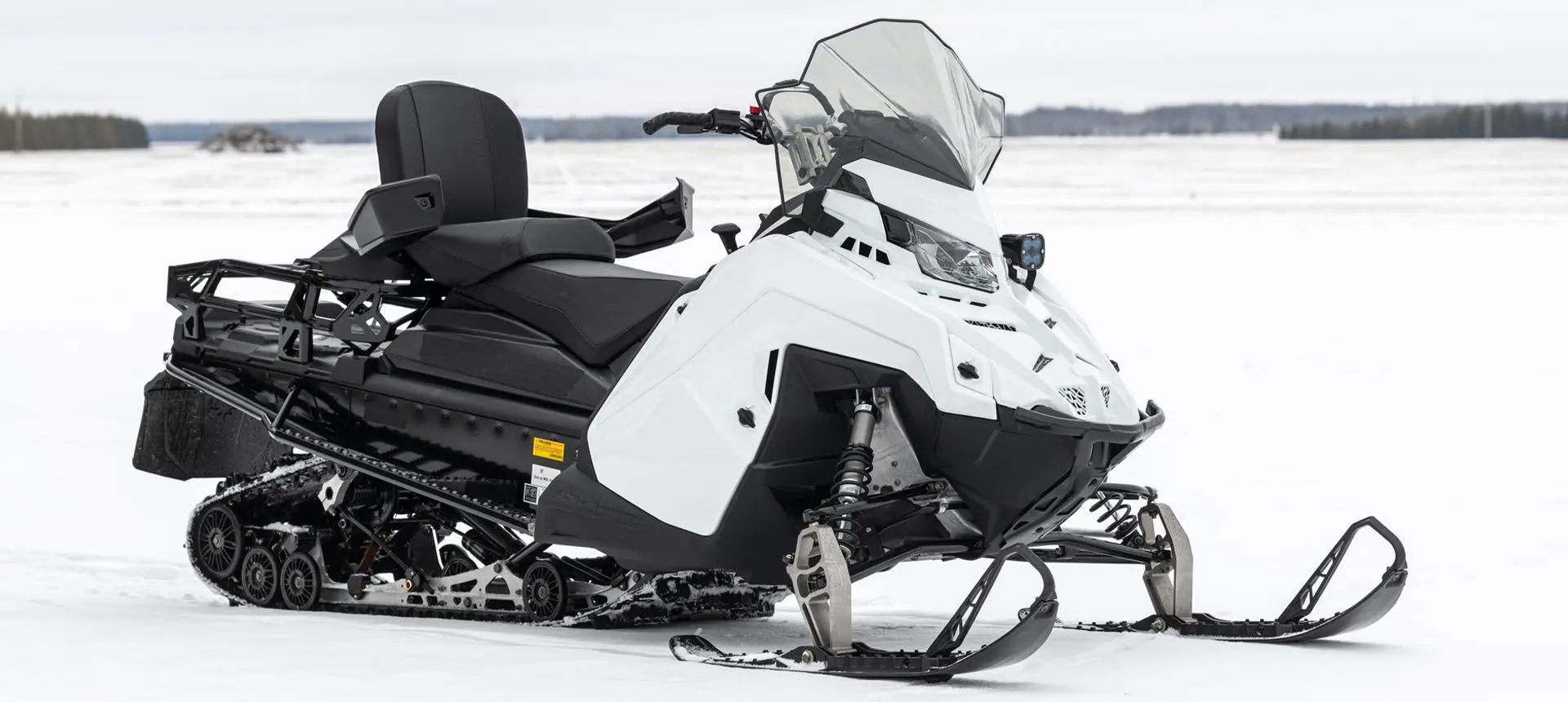 A white and black snowmobile 2026 Military 850 TITAN 155 sits on a snow-covered plain against a distant treeline.