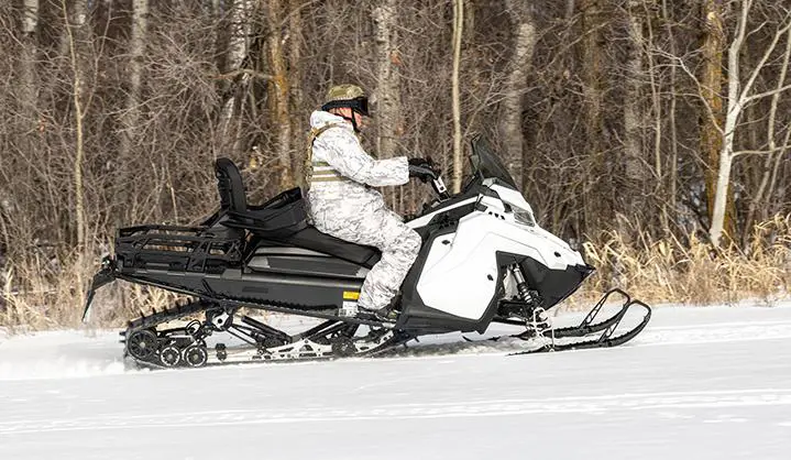 A person dressed in white camouflage drives a white and black snowmobile 2026 Military 850 TITAN 155 across a snow-covered field with trees in the background.