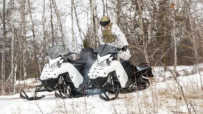 A person wearing winter camouflage stands between two white snowmobiles 2026 Military 850 TITAN 155 in a snowy, wooded area.