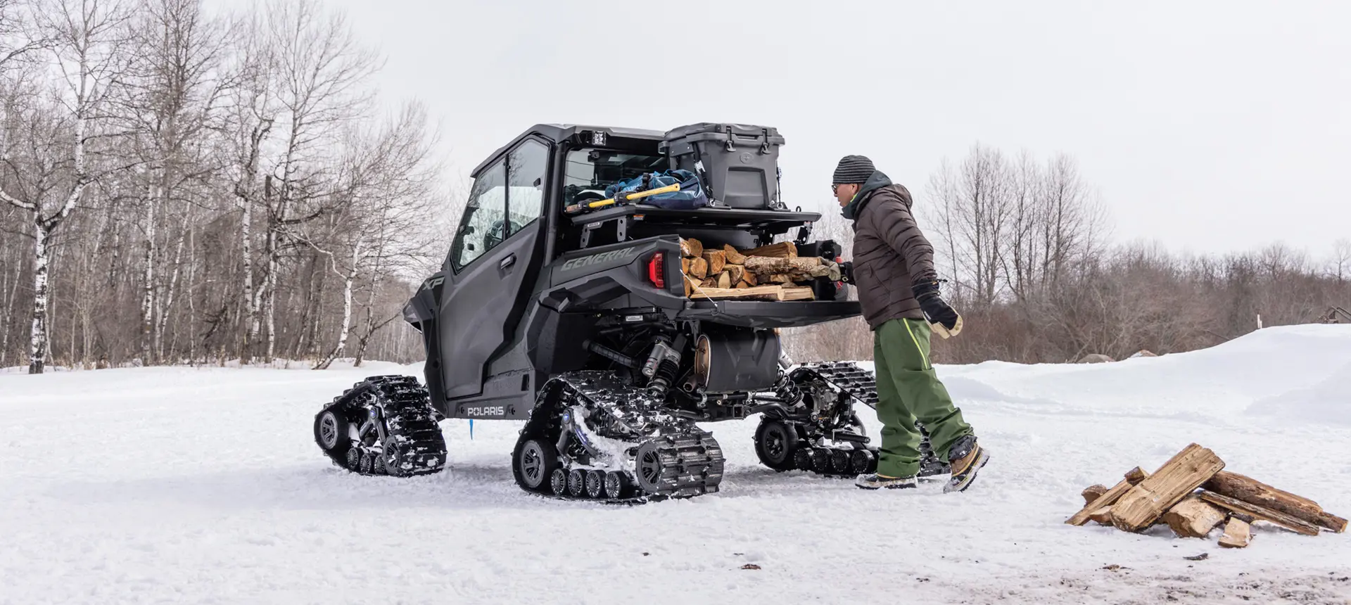 A person loading firewood into the cargo box of a winterized Polaris GENERAL XP 1000 side-by-side in the winter.