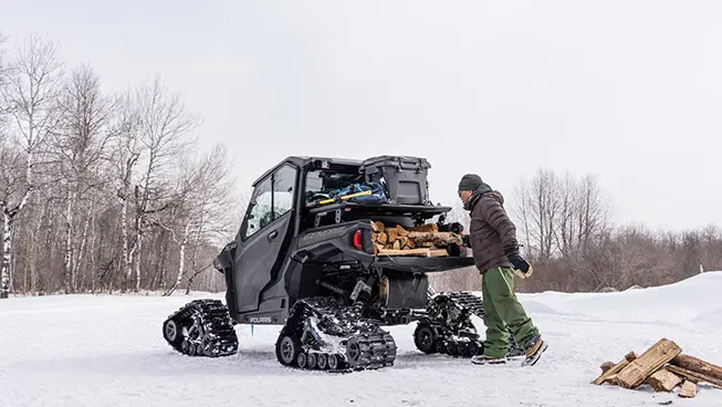 A person loading firewood into the cargo box of a winterized Polaris GENERAL XP 1000 side-by-side in the winter.