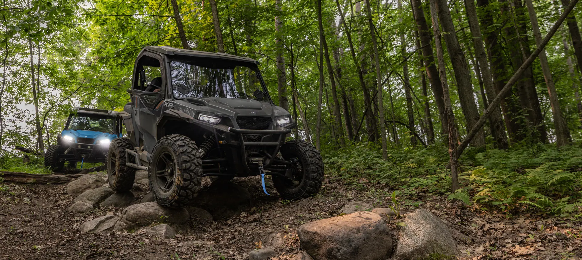 2 Polaris GENERAL XP 1000 side-by-sides driving down a forest trail and traversing rocky terrain.