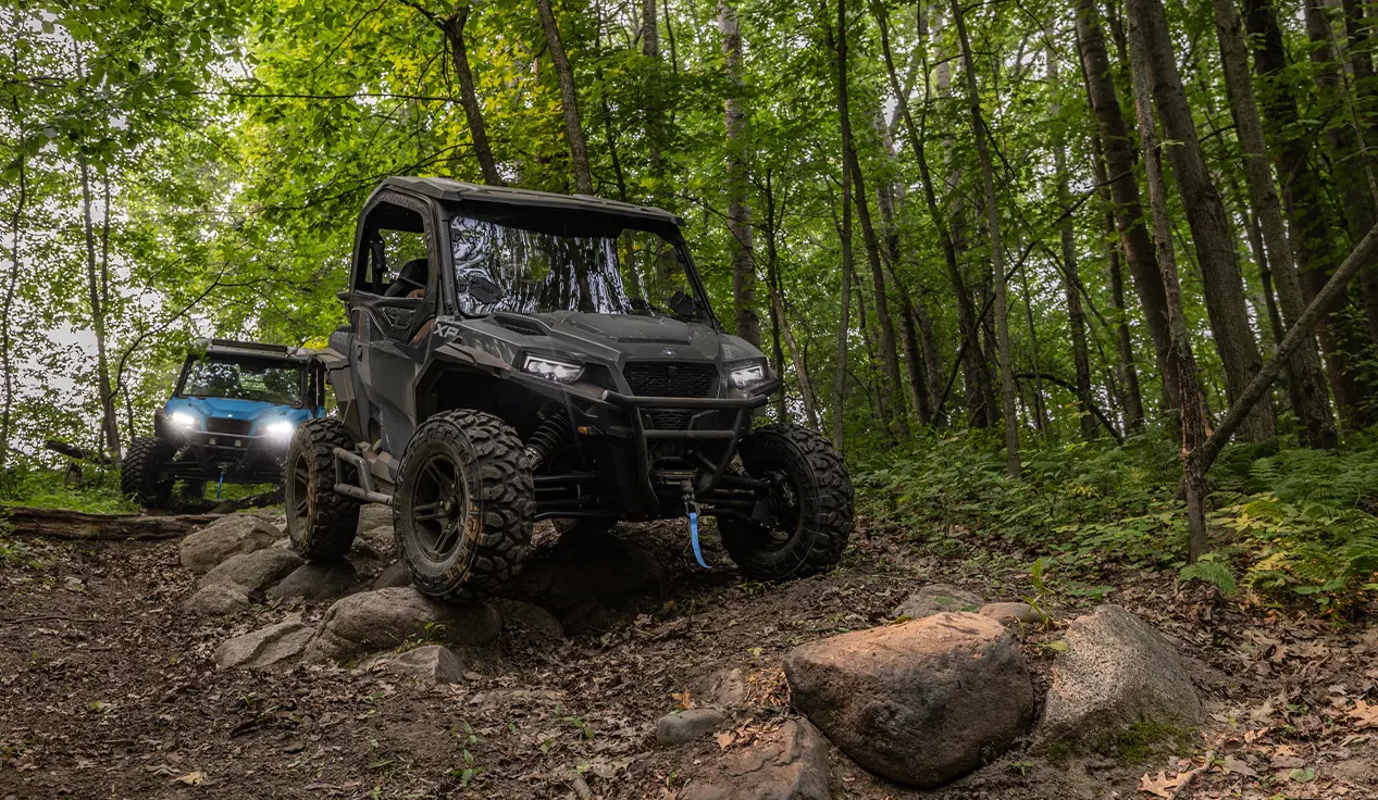 2 Polaris GENERAL XP 1000 side-by-sides driving down a forest trail and traversing rocky terrain.