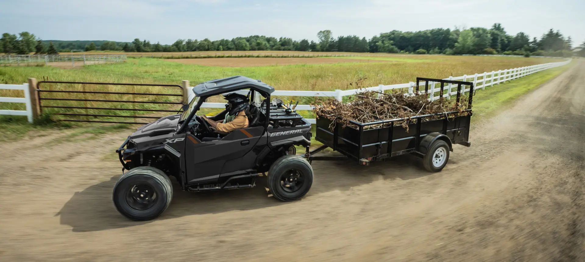 2 riders in a Polaris GENERAL XP 1000 side-by-side driving by a farm and pulling a trailer loaded with branches.