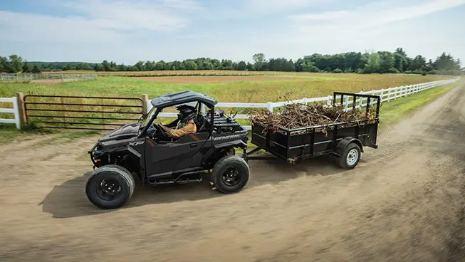 2 riders in a Polaris GENERAL XP 1000 side-by-side driving by a farm and pulling a trailer loaded with branches.
