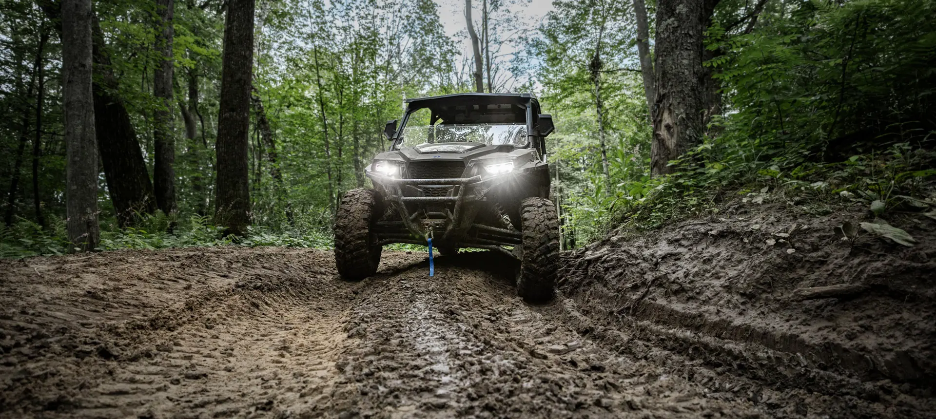 Front end view of a Polaris GENERAL XP 1000 side-by-side with headlights illuminated driving on a muddy trail.