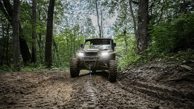 Front end view of a Polaris GENERAL XP 1000 side-by-side with headlights illuminated driving on a muddy trail.