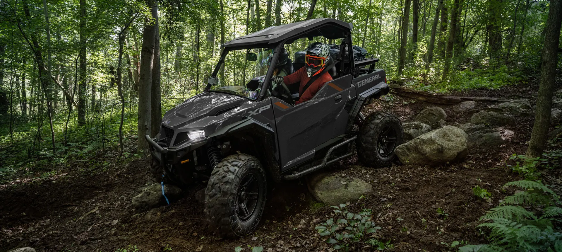 Side view of a Polaris GENERAL XP 1000 side-by-side with headlights illuminated driving over rocky terrain on a forest trail.