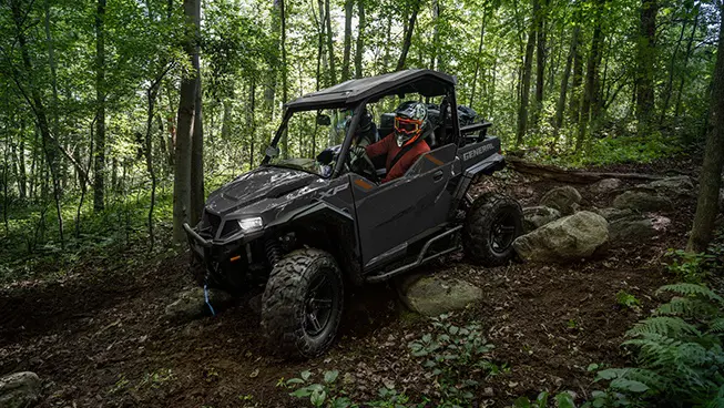 Side view of a Polaris GENERAL XP 1000 side-by-side with headlights illuminated driving over rocky terrain on a forest trail.