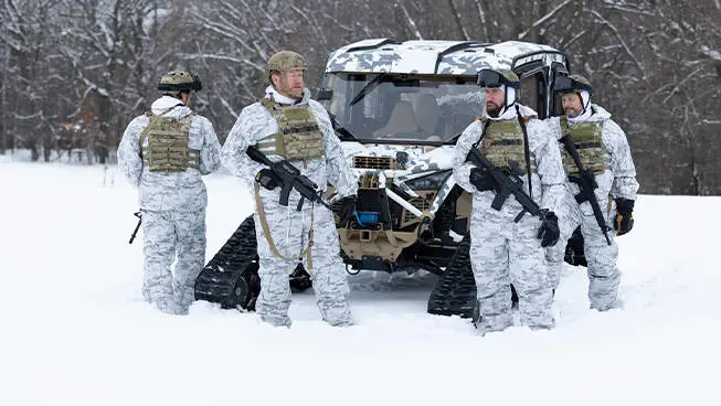 4 soldiers in winter garb standing near a MRZR Alpha military tactical side-by-side with tracks installed.