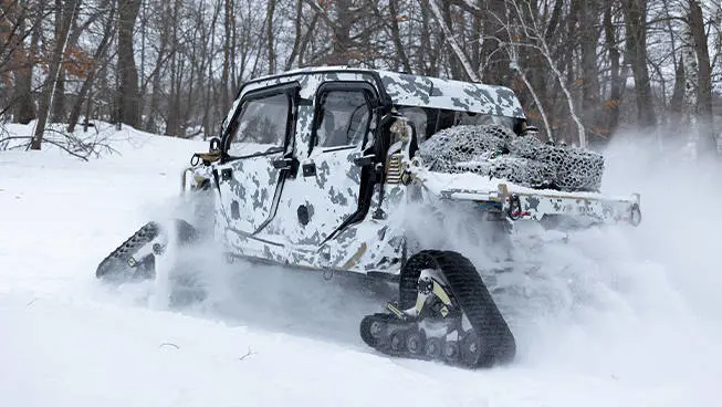 Side profile of a MRZR Alpha XP military tactical side-by-side with tracks installed driving on a snowy trail.