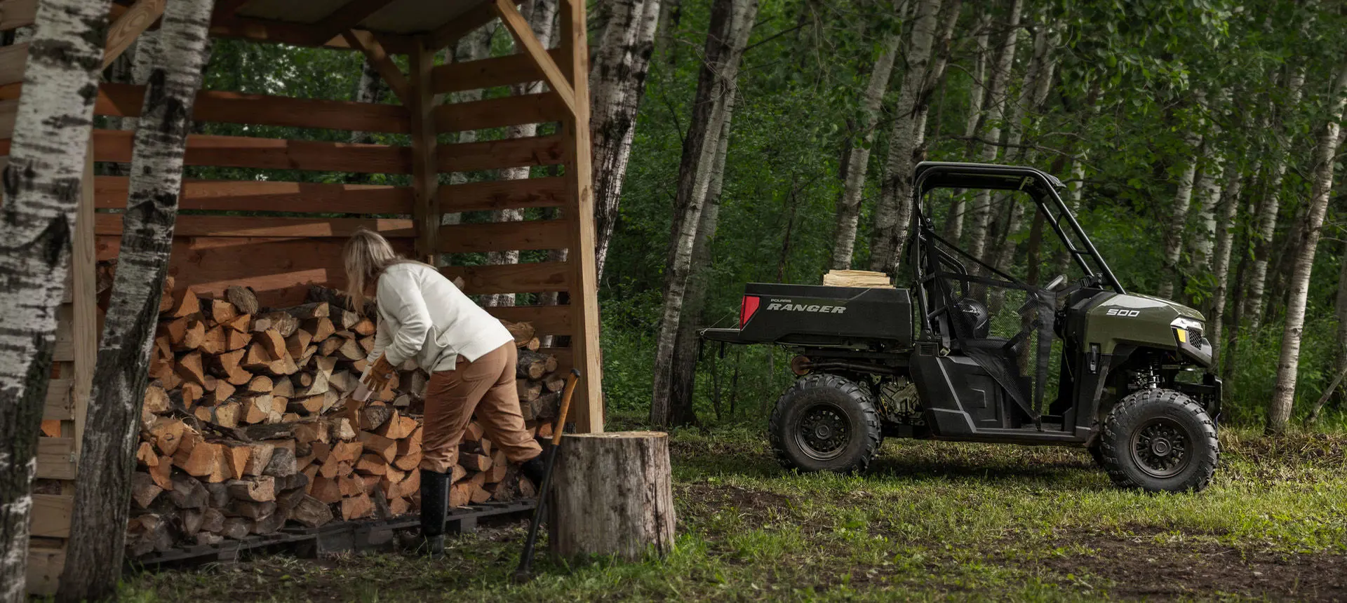 A woman splitting wood and loading it into the cargo box of a Sage Green Polaris RANGER 500 UTV.