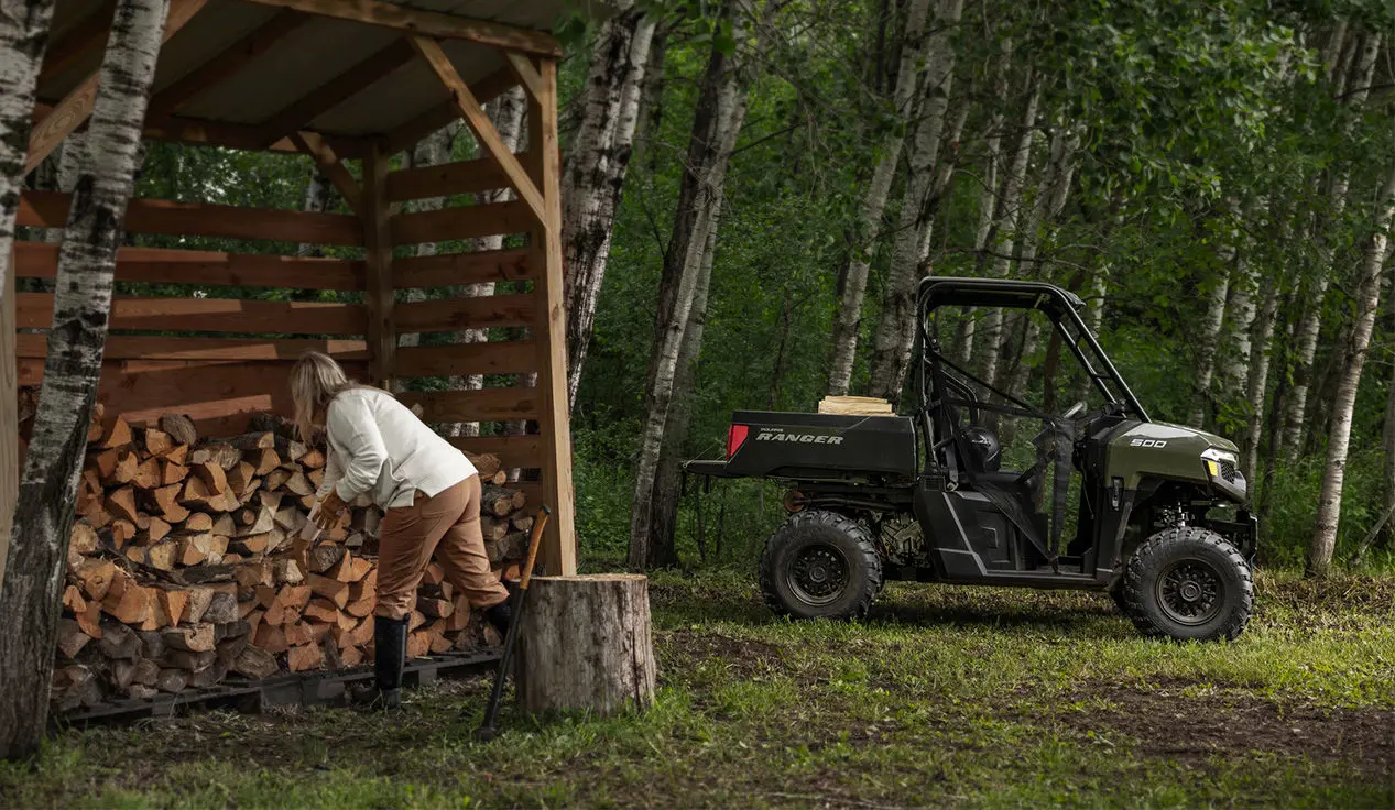 A woman splitting wood and loading it into the cargo box of a Sage Green Polaris RANGER 500 UTV.