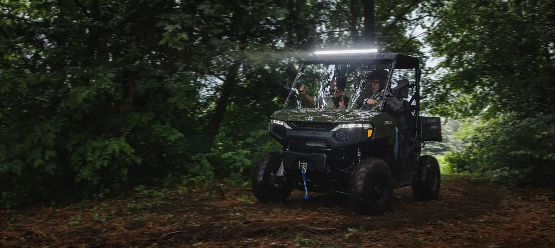 2 riders in a Sage Green Polaris RANGER 500 UTV with lightbar illuminated driving in the forest.