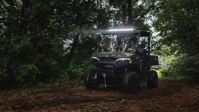 2 riders in a Sage Green Polaris RANGER 500 UTV with lightbar illuminated driving in the forest.