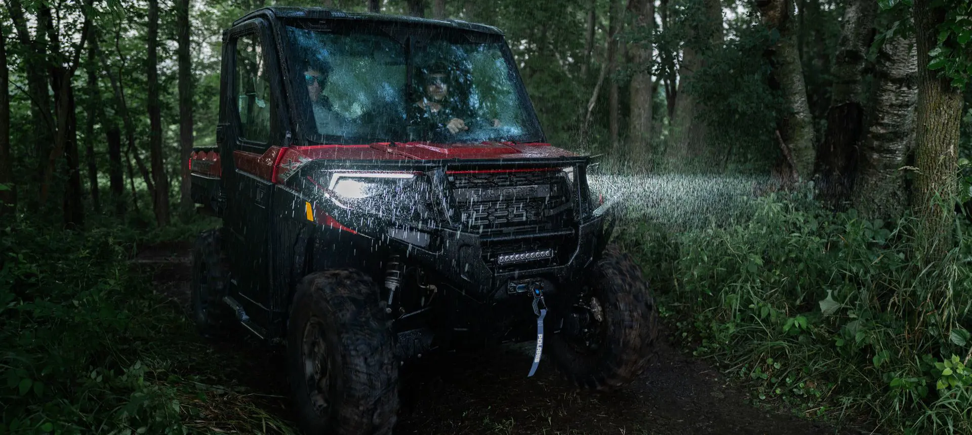 A Polaris RANGER XP 1000 NorthStar Edition UTV driving in the rain in a forest with headlights illuminated.