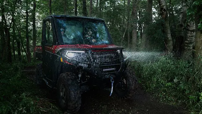 A Polaris RANGER XP 1000 NorthStar Edition UTV driving in the rain in a forest with headlights illuminated.