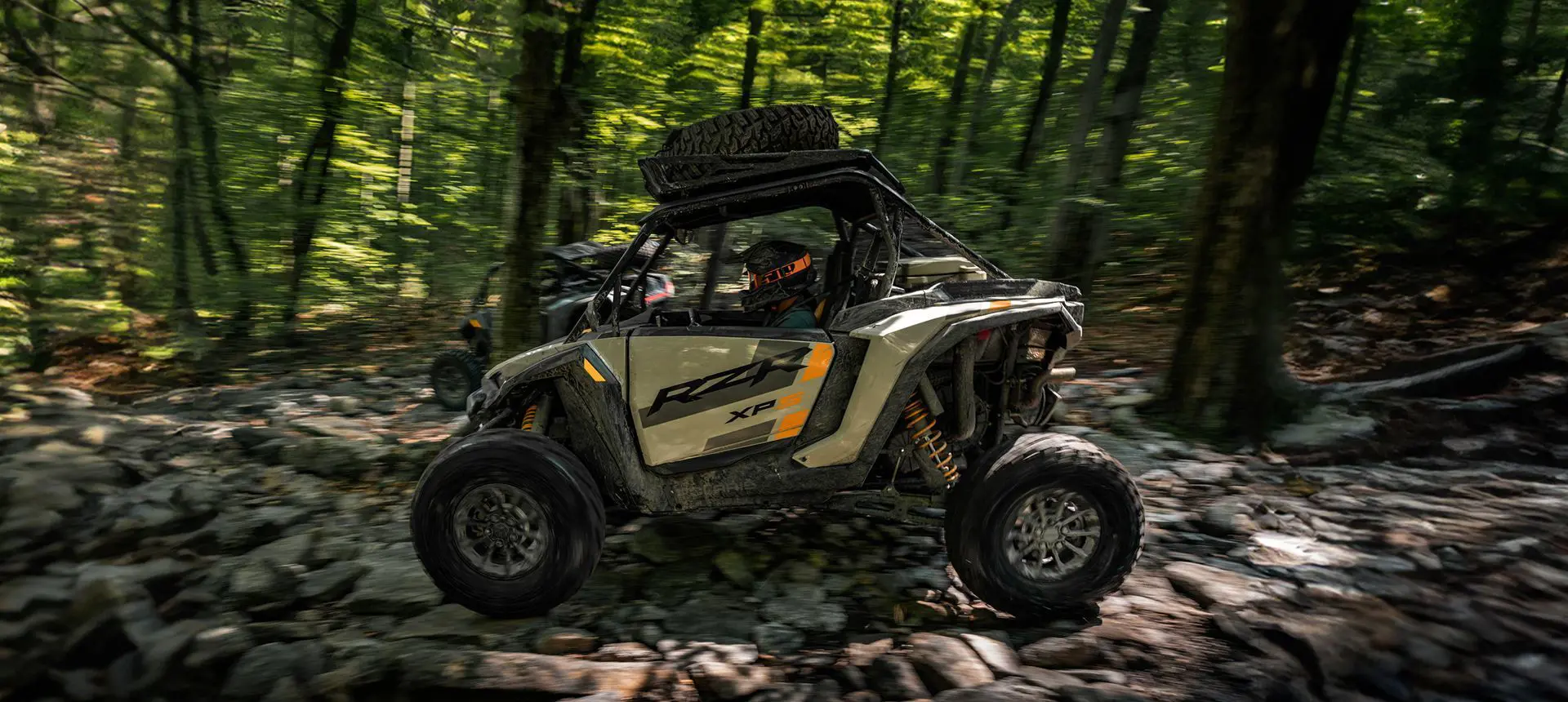 Side profile of a RZR XP S side-by-side driving over rocky terrain in the forest.