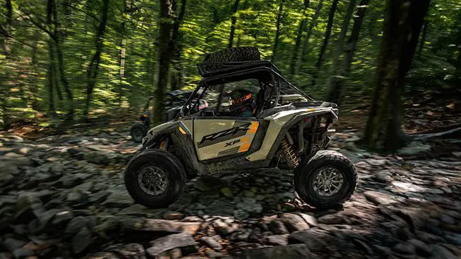 Side profile of a RZR XP S side-by-side driving over rocky terrain in the forest.