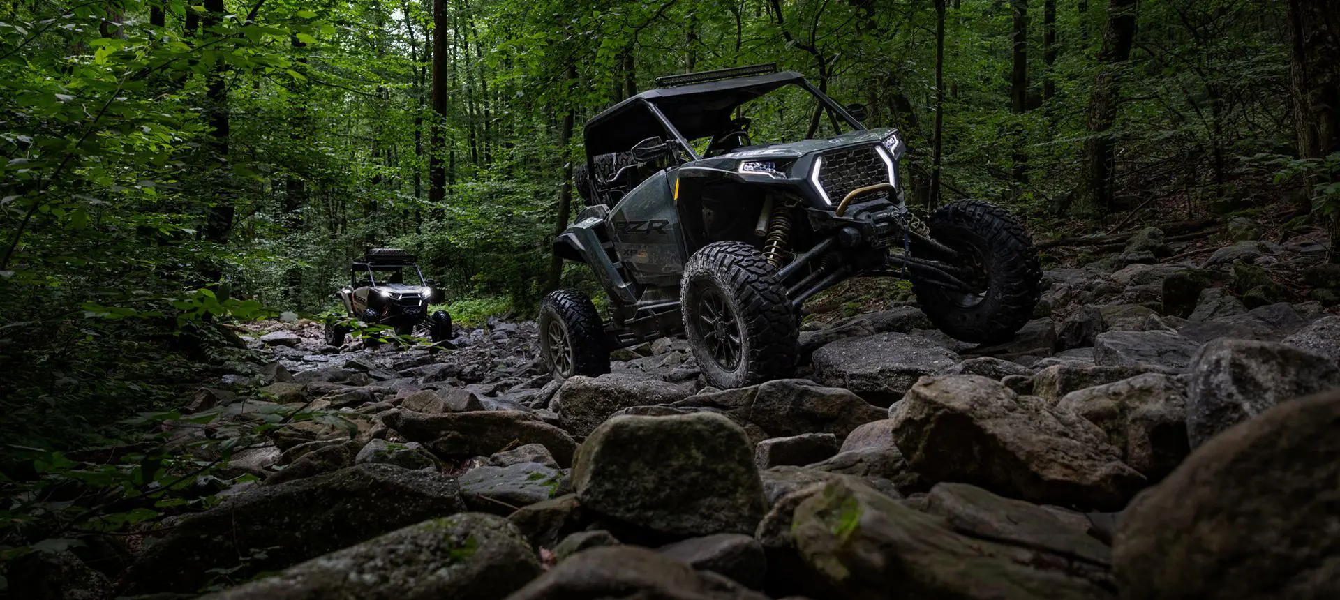 2 RZR XP side-by-sides driving over rocky terrain in the forest.