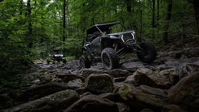 2 RZR XP side-by-sides driving over rocky terrain in the forest.