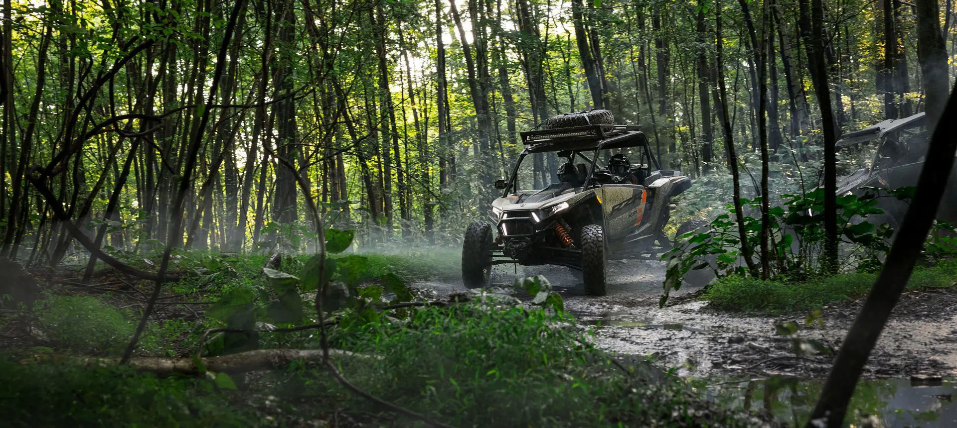 A muddy RZR XP S side-by-side driving through the forest.
