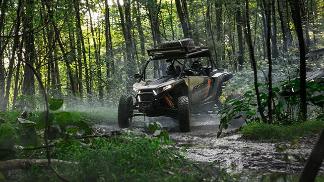 A muddy RZR XP S side-by-side driving through the forest.
