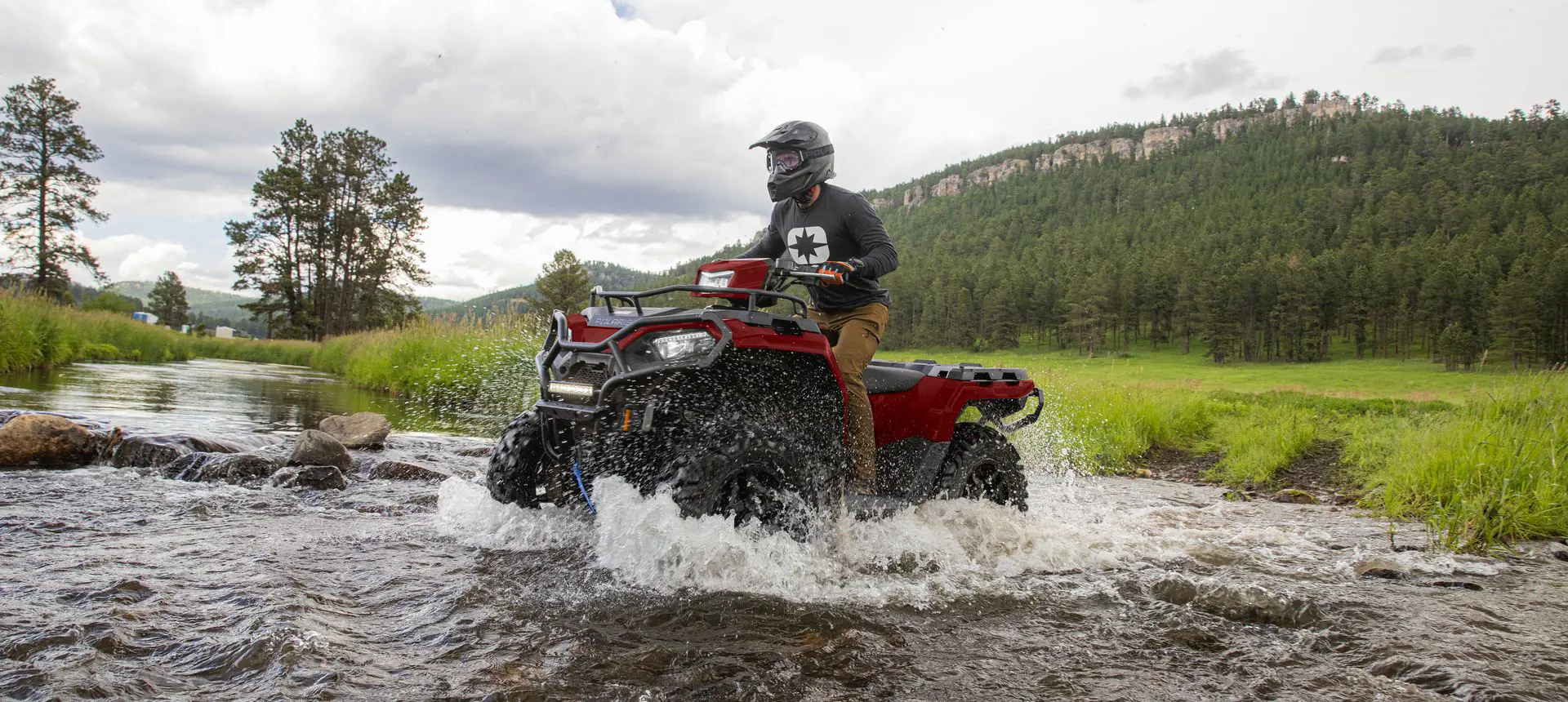 A rider on a Polaris Sportsman 570 ATV driving through a shallow riverbed.