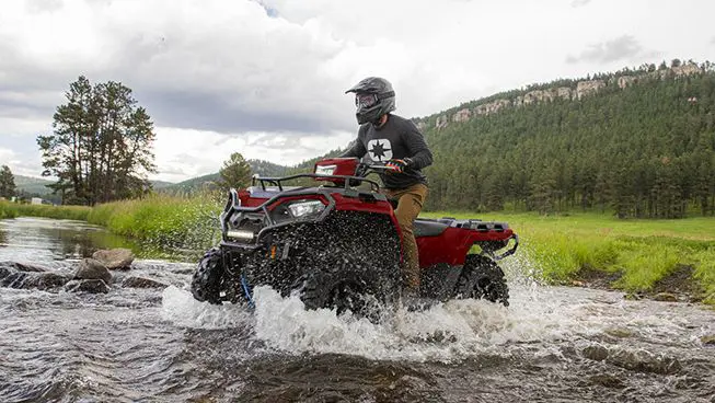 A rider on a Polaris Sportsman 570 ATV driving through a shallow riverbed.