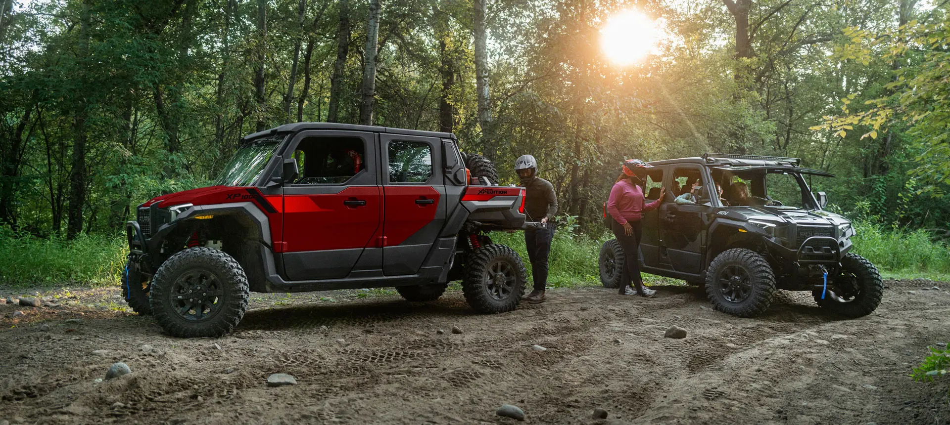 A performance red Polaris XPEDITION XP 5 NorthStar side-by-side and a Matte Super Graphite Polaris XPEDITION XP 5 NorthStar side-by-side parked with drivers preparing for a ride.