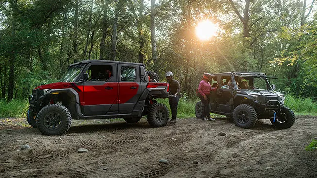 A performance red Polaris XPEDITION XP 5 NorthStar side-by-side and a Matte Super Graphite Polaris XPEDITION XP 5 NorthStar side-by-side parked with drivers preparing for a ride.