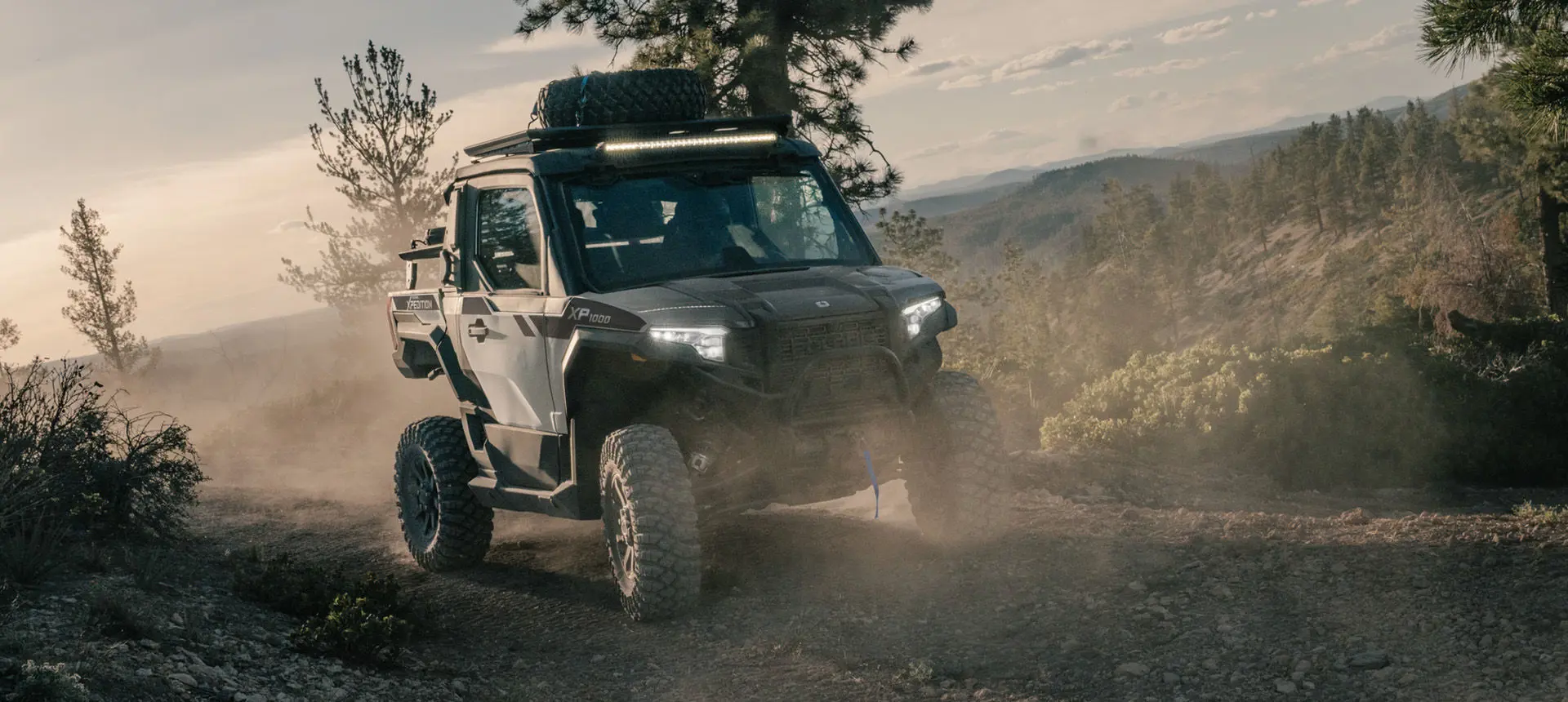 Front-end view of a Slate Gray Polaris XPEDITION XP NorthStar side-by-side with headlights illuminated on a trail.
