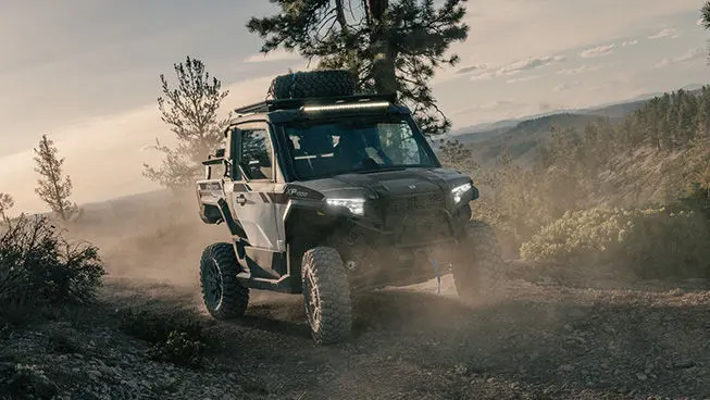 Front-end view of a Slate Gray Polaris XPEDITION XP NorthStar side-by-side with headlights illuminated on a trail.
