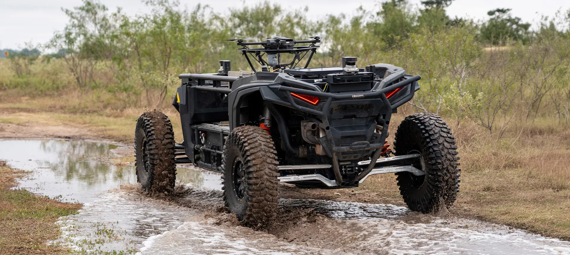Rear view of a black Polaris RZR XP 1000 equipped with sensors and a drone, splashing through shallow water.