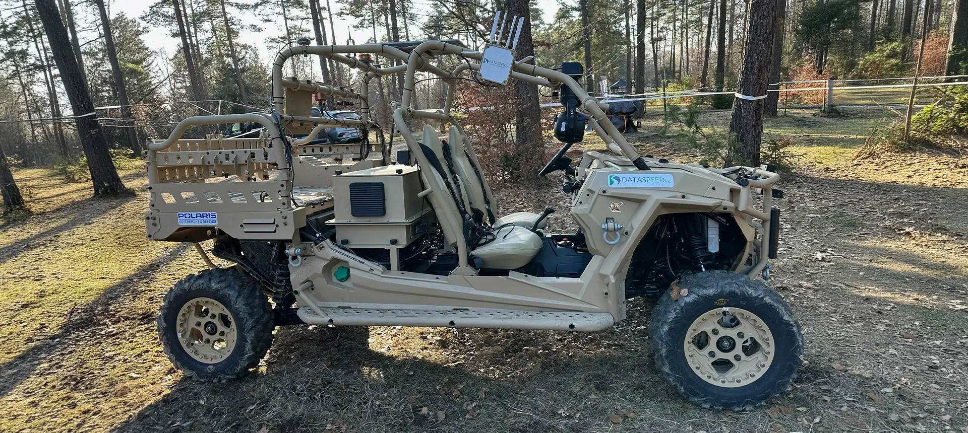 Side profile of a tan Polaris MRZR D4 ultralight tactical vehicle equipped with sensor technology, parked in a forest.