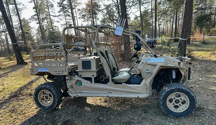 Side profile of a tan Polaris MRZR D4 ultralight tactical vehicle equipped with sensor technology, parked in a forest.