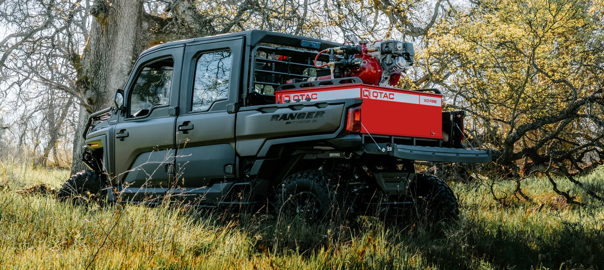 Polaris RANGER CREW XD 1500 NorthStar Edition parked in tall grass with a QTAC firefighting unit mounted in the rear bed, surrounded by trees in a wooded outdoor setting.