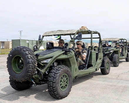 Polaris MRZR military SxS vehicles lined up on a paved surface, with personnel seated inside and additional units visible in formation behind, showing mounted spare tires and tactical configurations.