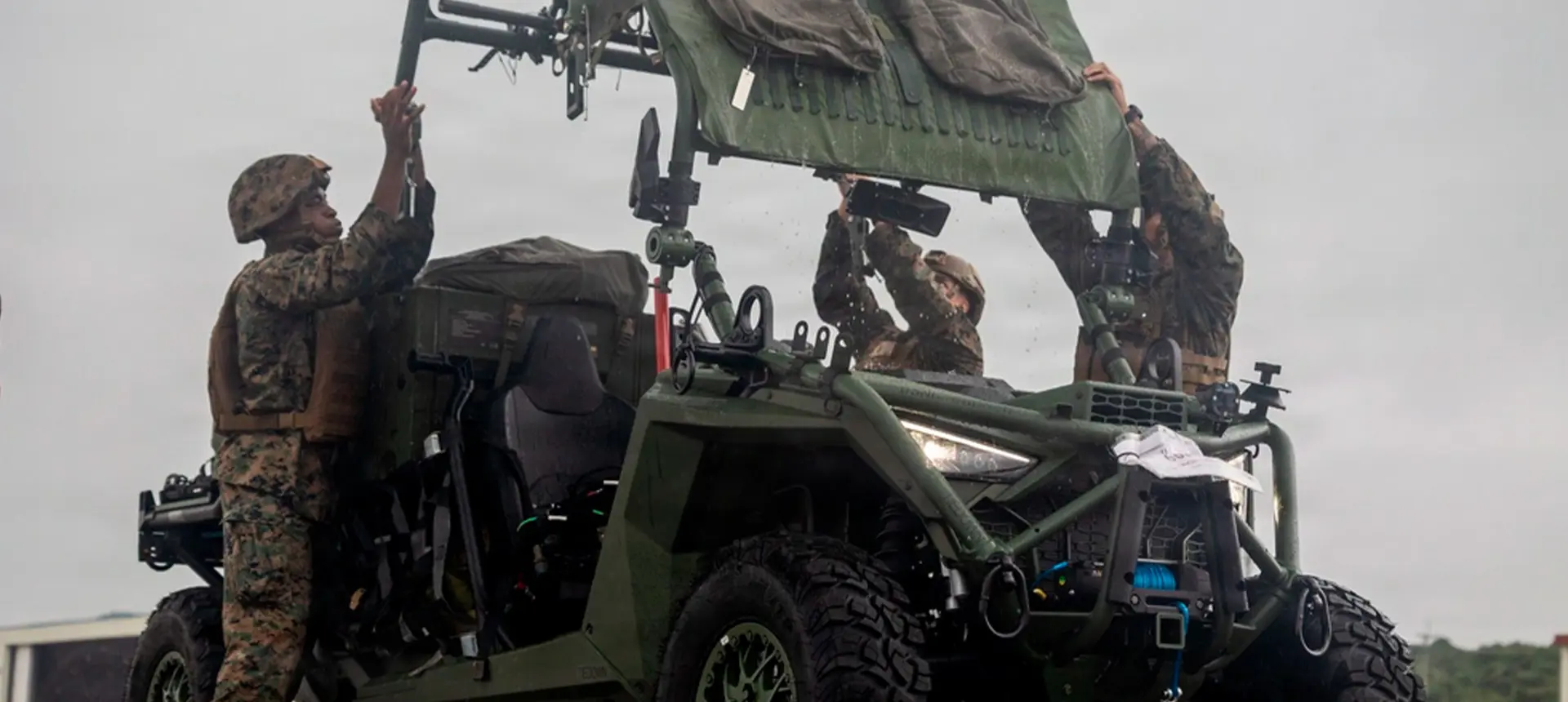 Military personnel loading equipment onto a Polaris MRZR military Side-by-Side vehicle in wet outdoor conditions with tactical gear mounted throughout the vehicle.