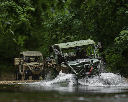 Polaris MRZR military SxS vehicles lined up on a paved surface, with personnel seated inside and additional units visible in formation behind, showing mounted spare tires and tactical configurations.