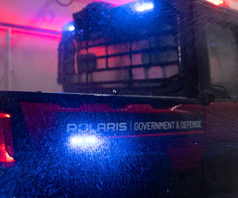 Polaris Government and Defense vehicle undergoing a water intrusion test in a controlled lab environment, exposed to heavy simulated rainfall with emergency lights activated.