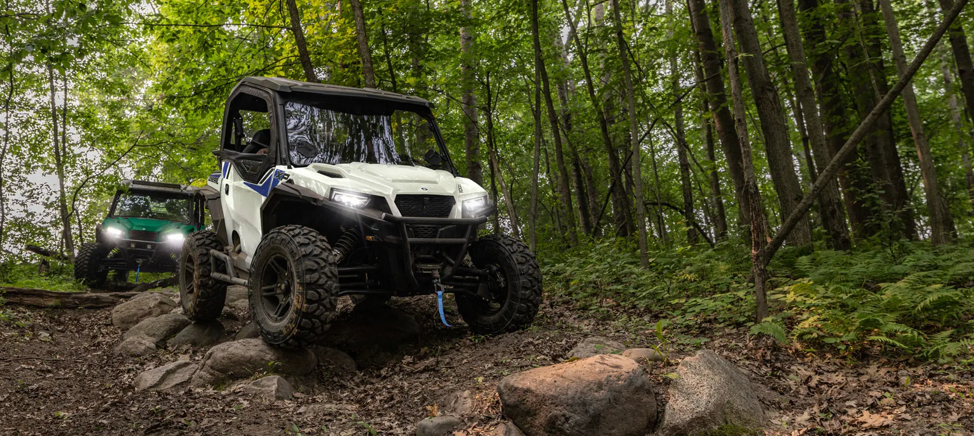 2 Polaris GENERAL XP 1000 side-by-sides driving down a forest trail and traversing rocky terrain.