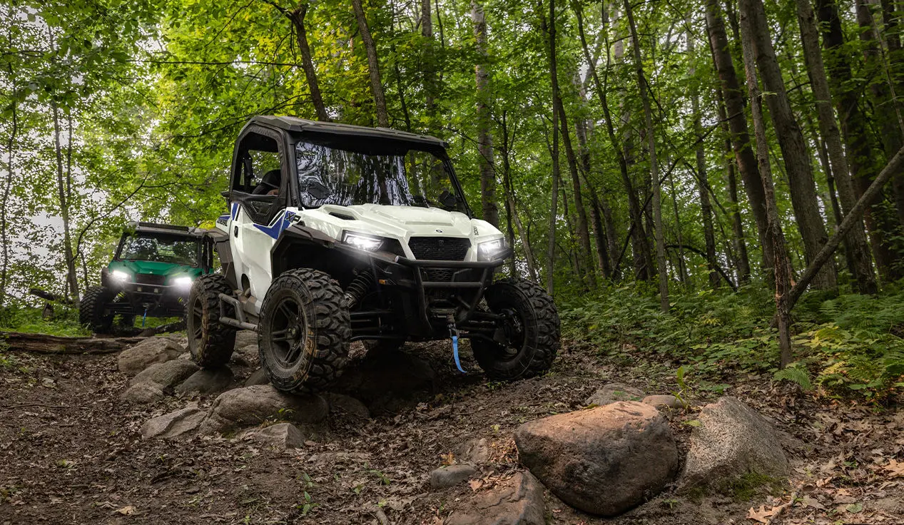 2 Polaris GENERAL XP 1000 side-by-sides driving down a forest trail and traversing rocky terrain.