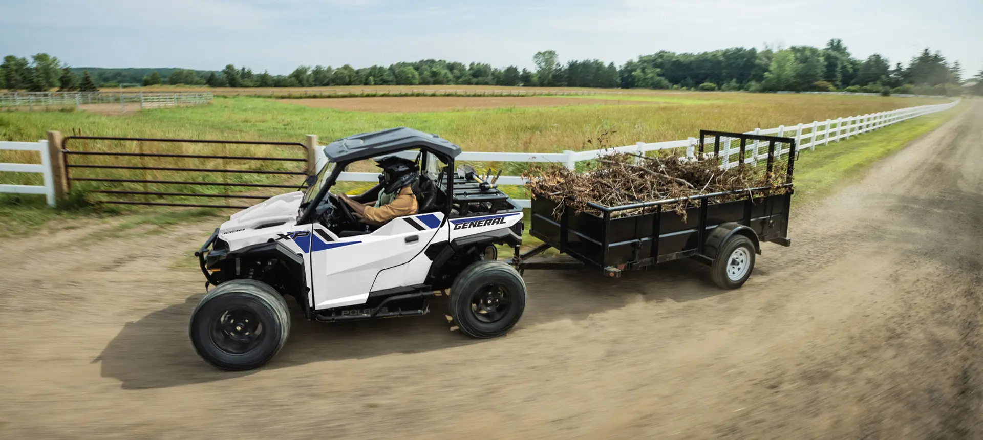 2 riders in a Polaris GENERAL XP 1000 side-by-side driving by a farm and pulling a trailer loaded with branches.