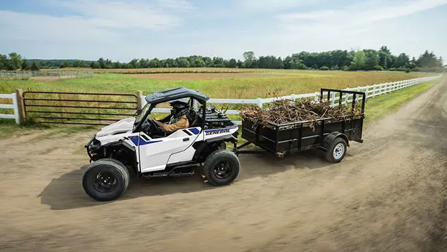 2 riders in a Polaris GENERAL XP 1000 side-by-side driving by a farm and pulling a trailer loaded with branches.